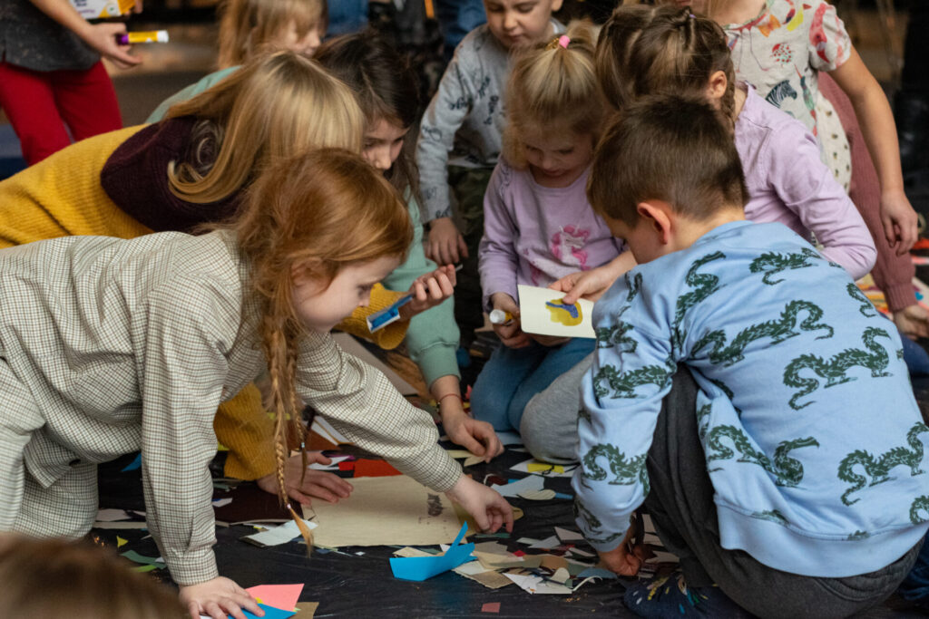 Dzieci siedzą wokół kolorowych papierowych figur geometrycznych i wybierają je do prac plastycznych.