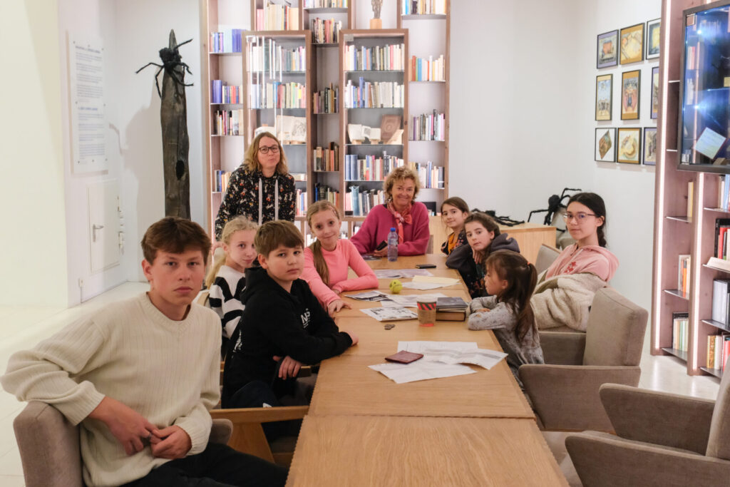 Uczestnicy i prowadzący siedzą przy stole w bibliotece i pozują do zdjęcia.