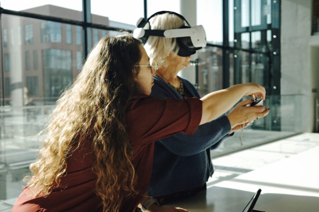 Opiekunka gry instruuje uczestniczkę ubraną w okulary VR jak używać kontrolera.